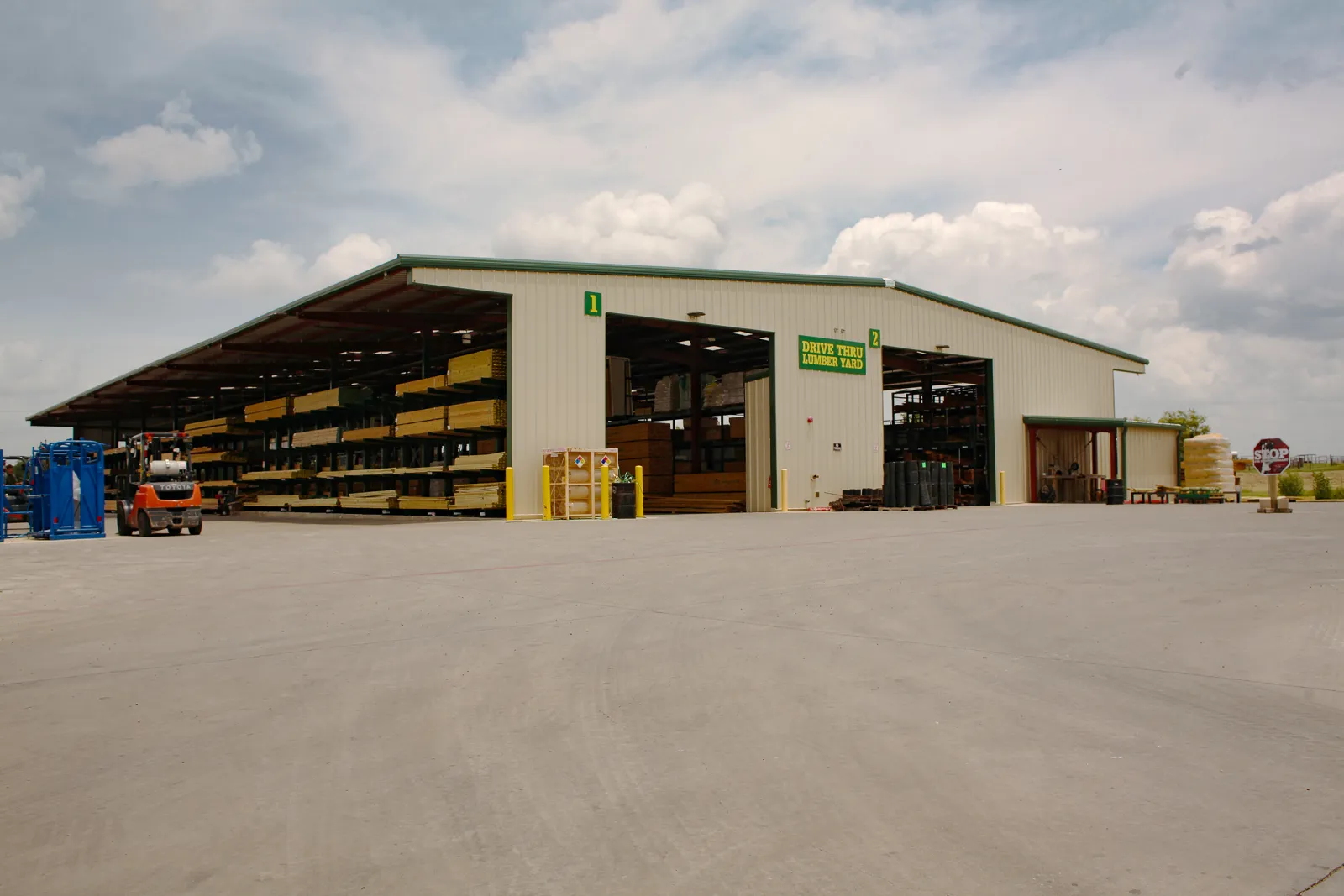 Large drive-thru lumber yard with stacks of wood, forklift, and clear sky above a concrete pavement.