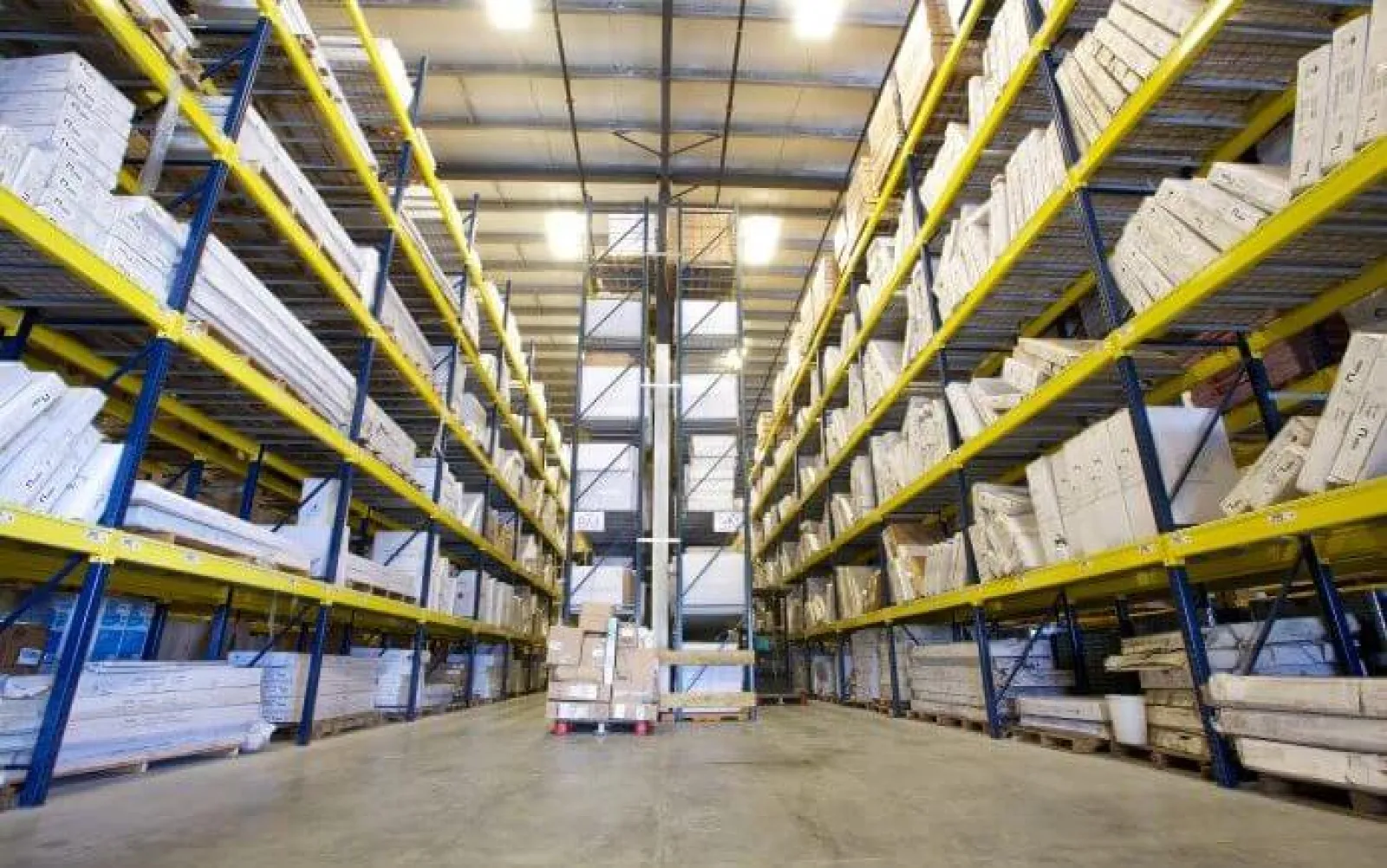 Large warehouse aisle with tall blue and yellow racks filled with stacked cardboard boxes and packages.