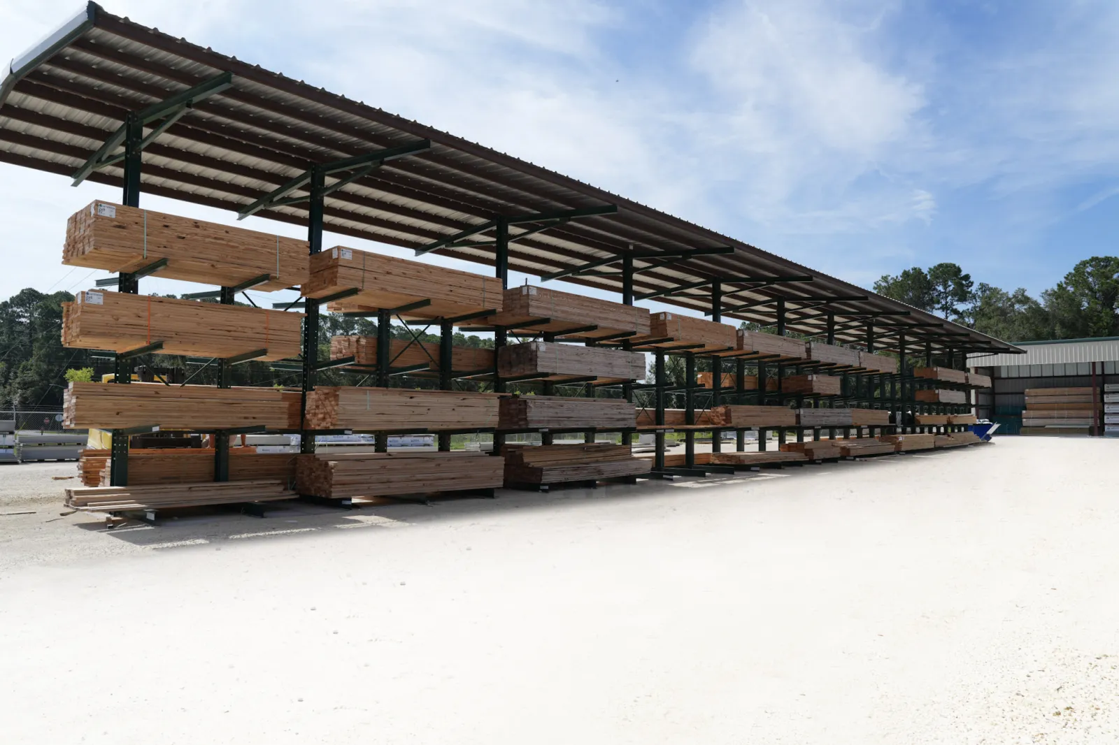 Outdoor lumber storage racks filled with wooden planks under a metal roof on a sunny day.