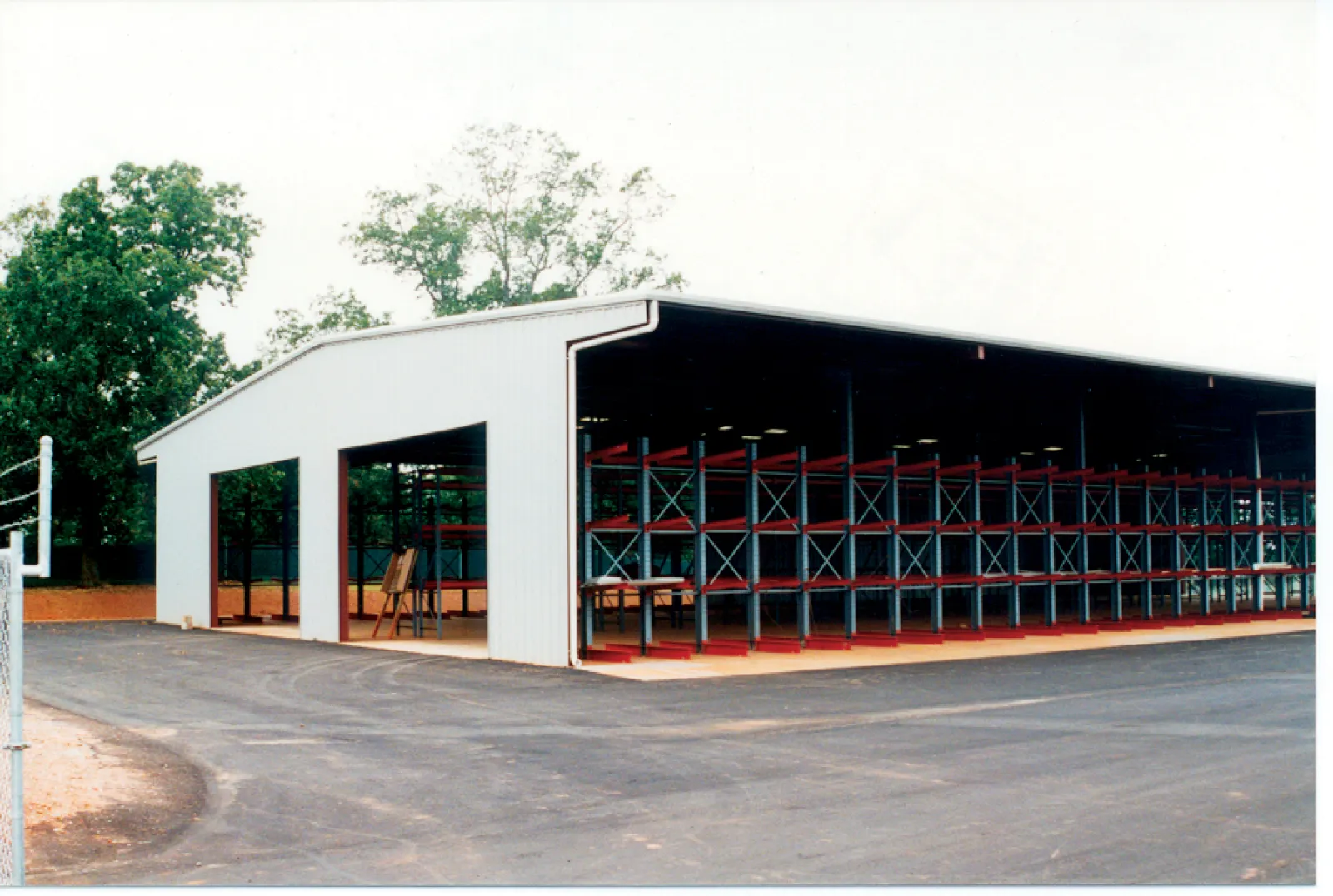 Large open warehouse with metal shelving units and paved driveway surrounded by trees on a clear day