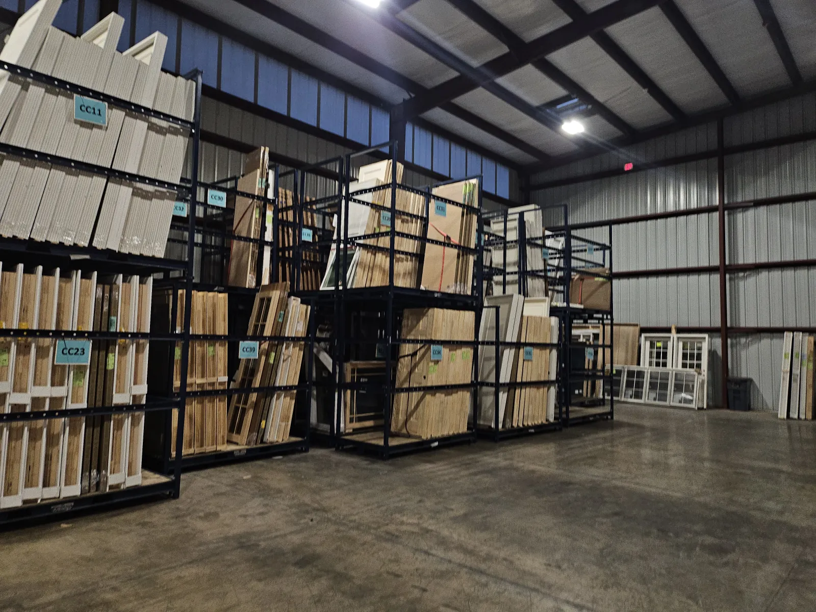 Warehouse interior with large metal racks storing flat wooden panels and frames under industrial lighting.
