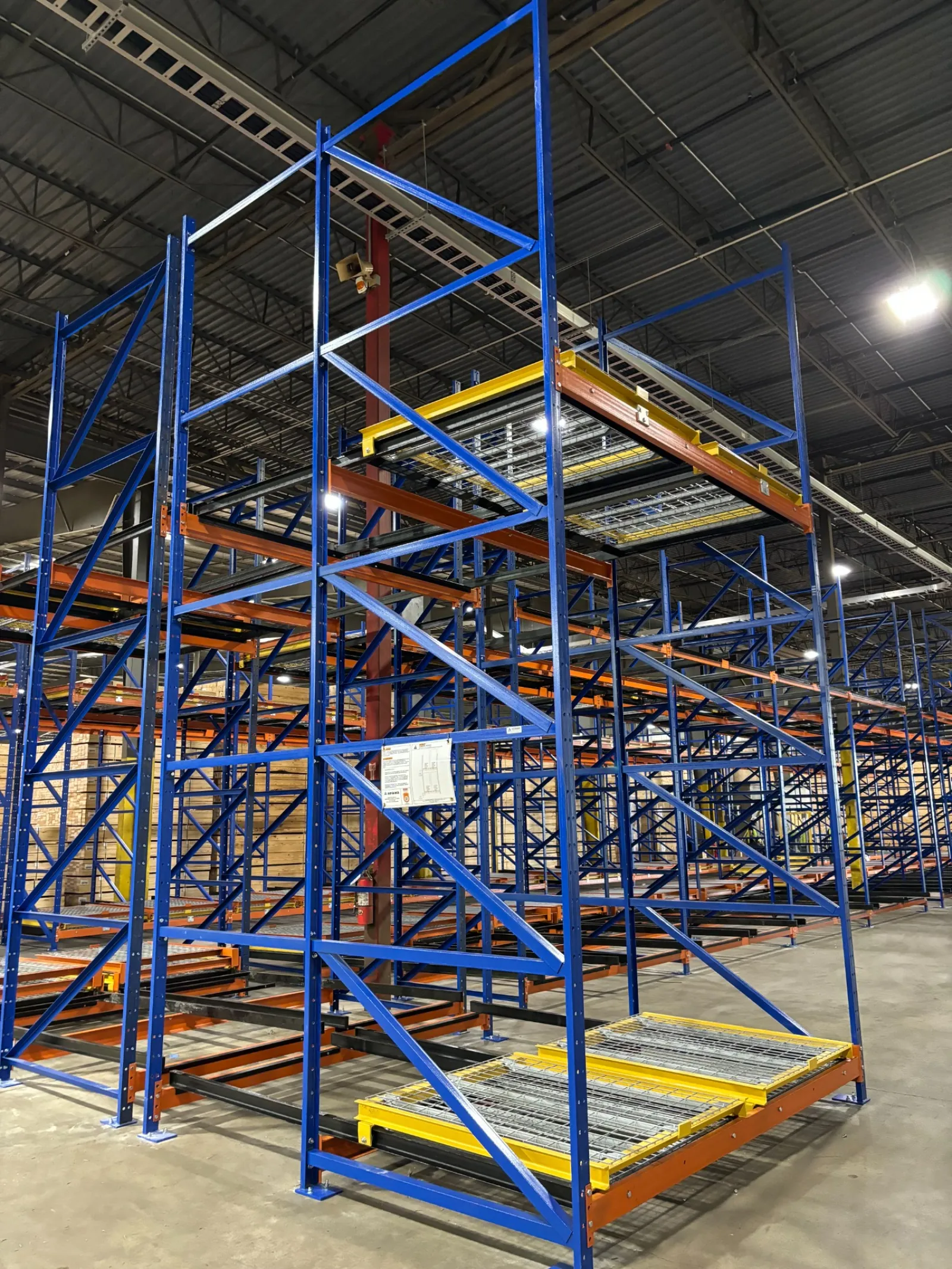 Empty blue and orange industrial warehouse shelving units under bright overhead lights on concrete floor.