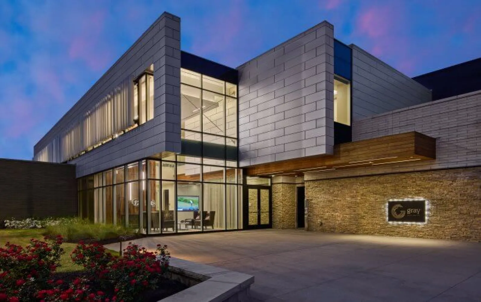 Modern commercial building with glass walls, stone facade, and illuminated interior at twilight under a purple sky.