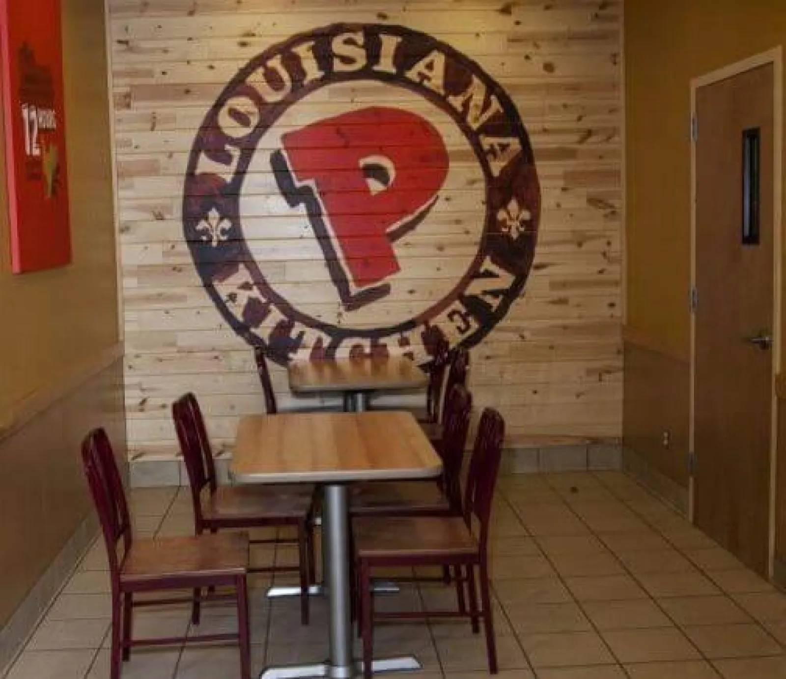 Interior of a Popeyes Louisiana Kitchen with wooden tables, chairs, and large logo mural on wood wall.