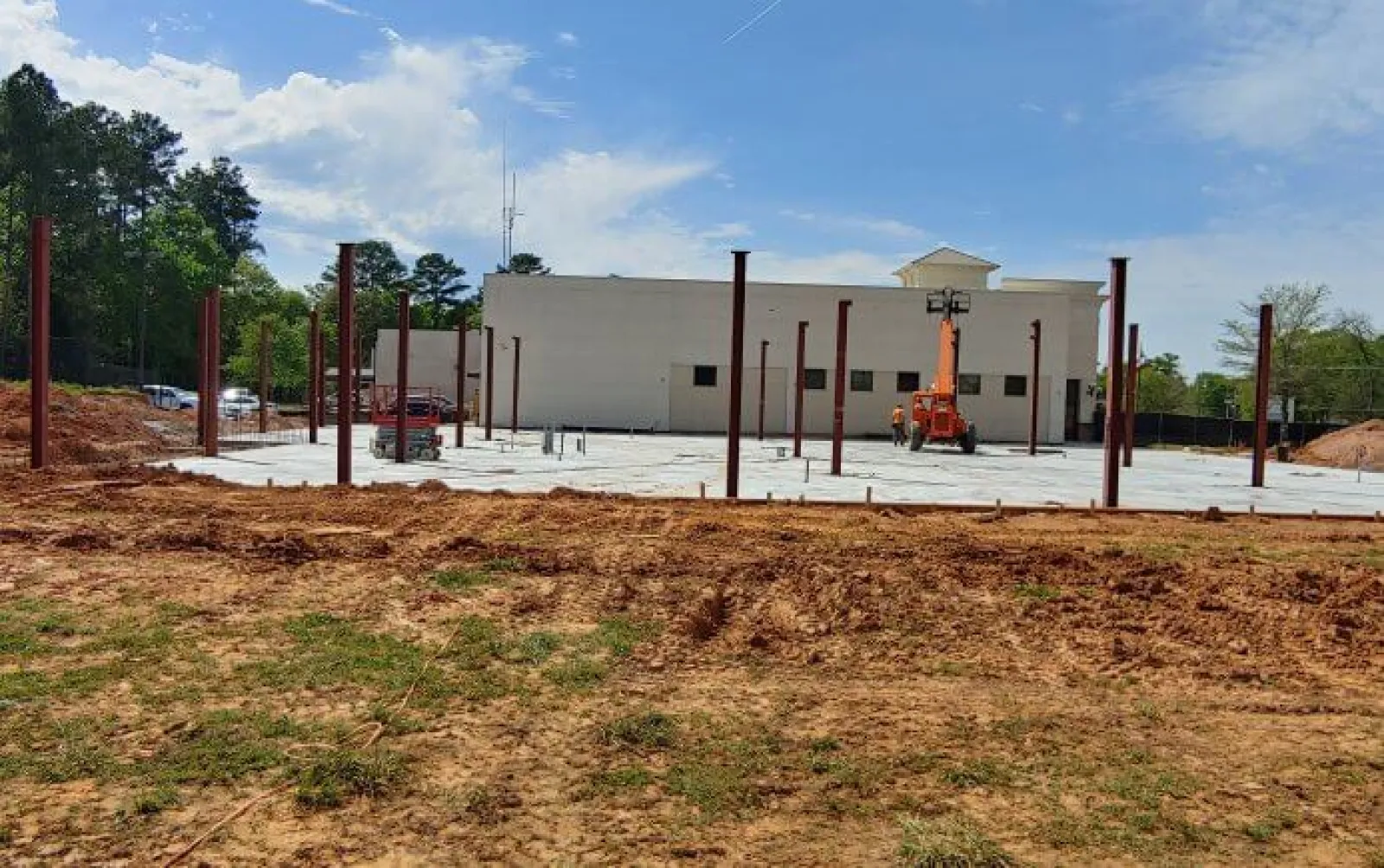 Construction site with steel beams installed on concrete foundation near a building under a blue sky.