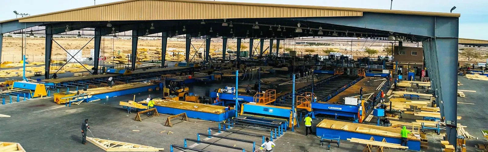 Wide view of an industrial metal fabrication workshop with workers and machinery under a large steel shed.