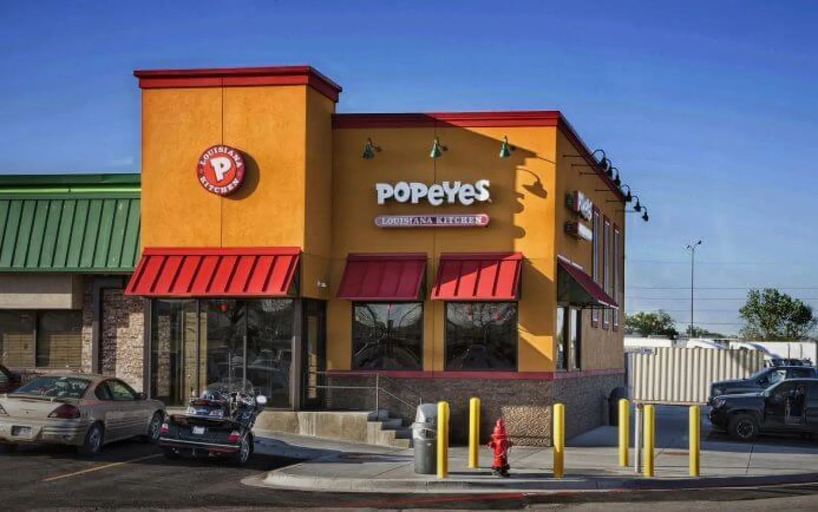 Exterior view of a Popeyes Louisiana Kitchen fast-food restaurant with bright orange and red facade under clear blue sky.