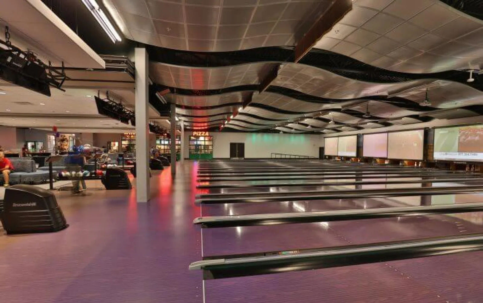 Interior view of a spacious bowling alley with multiple lanes, seating areas, and a pizza counter in the back.