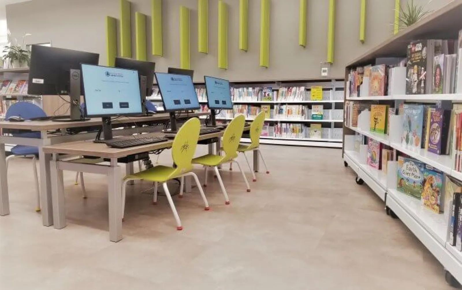 Library computer station with multiple monitors and green chairs beside bookshelves filled with books.