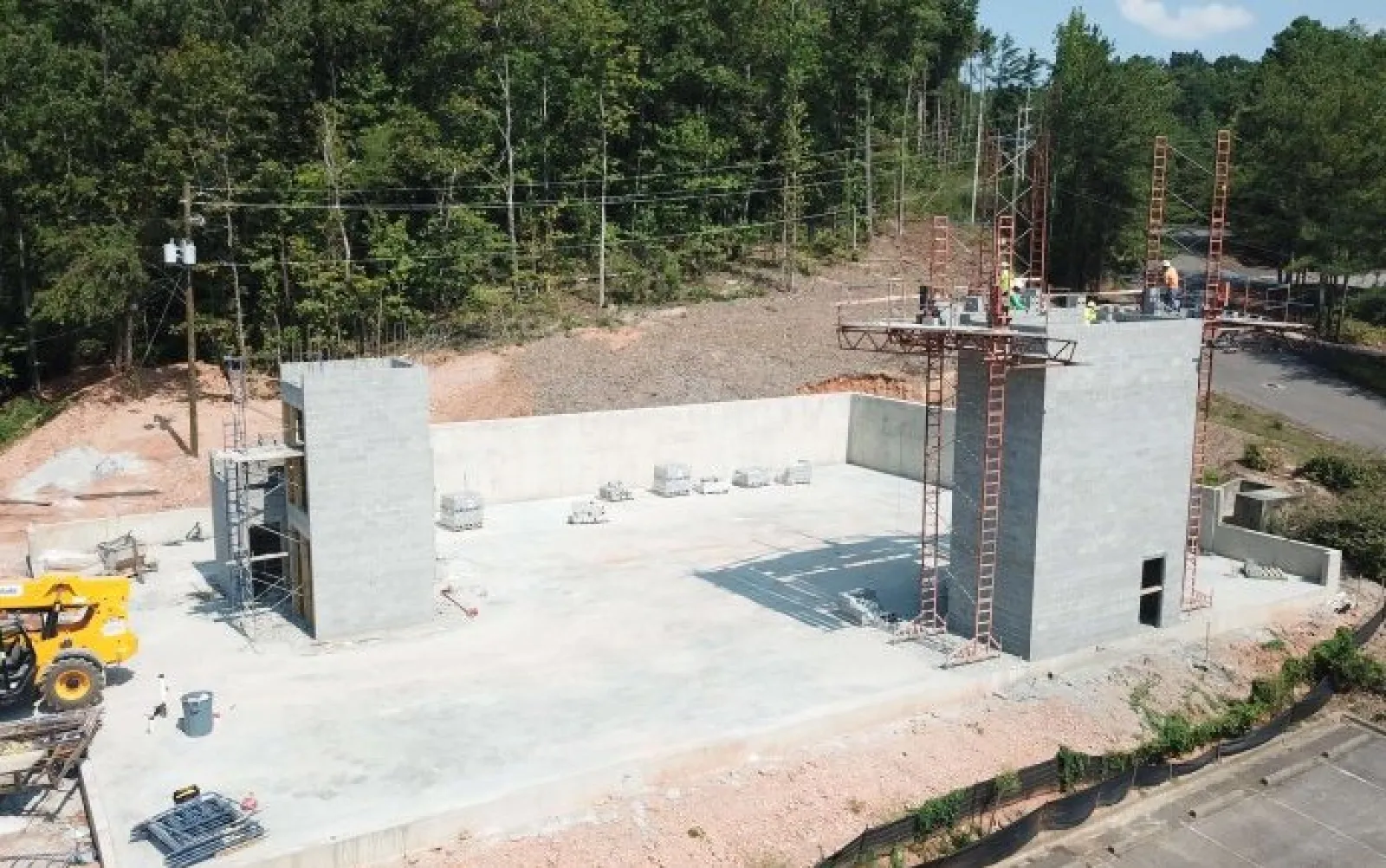 Construction site with concrete foundation and walls in early stages surrounded by trees and construction equipment.