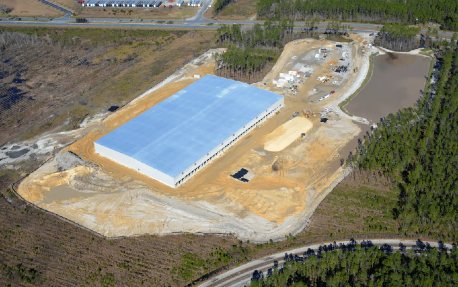 Aerial view of a large warehouse under construction surrounded by cleared land and forest with a nearby pond.