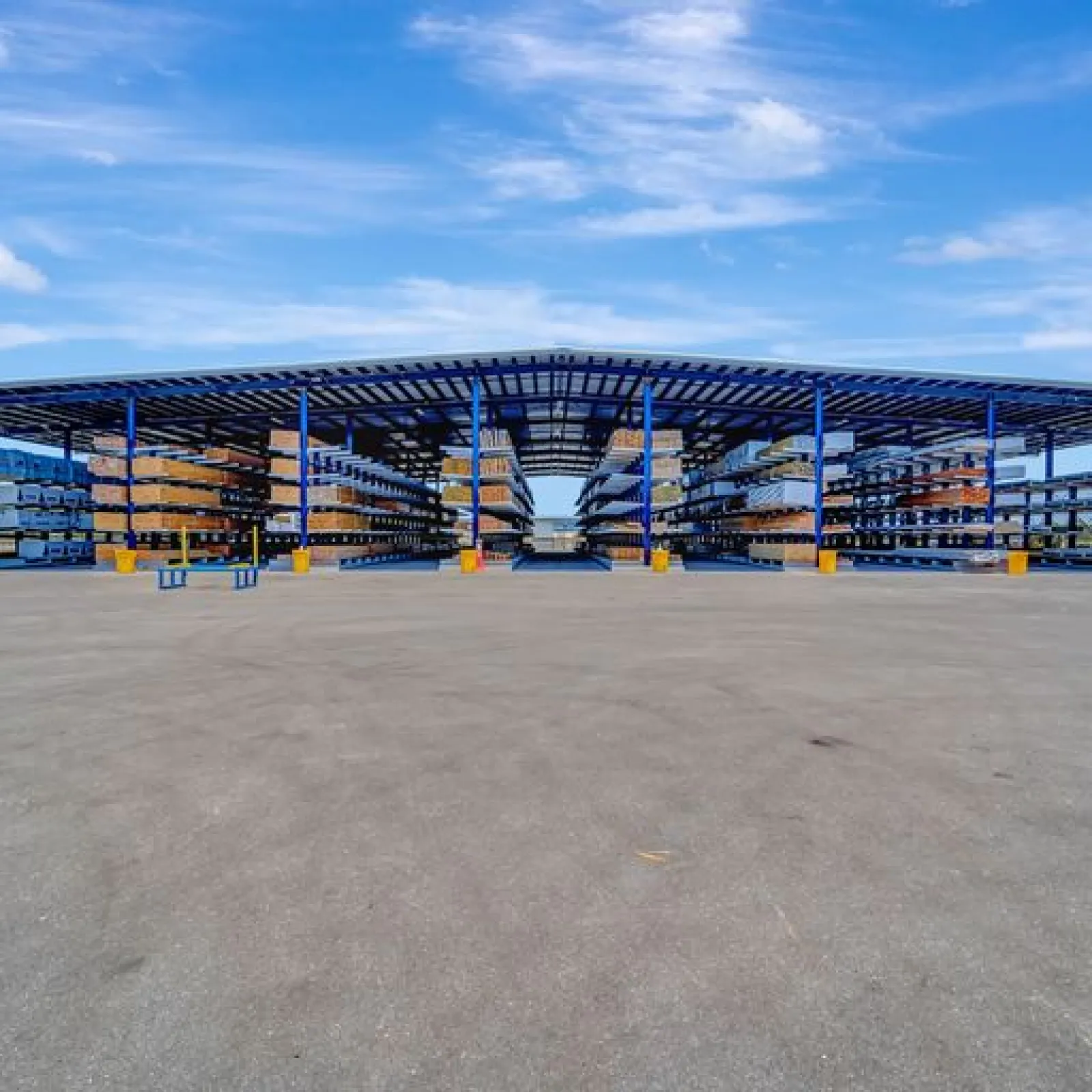 Large open warehouse with steel racks holding construction materials under a blue sky with clouds.