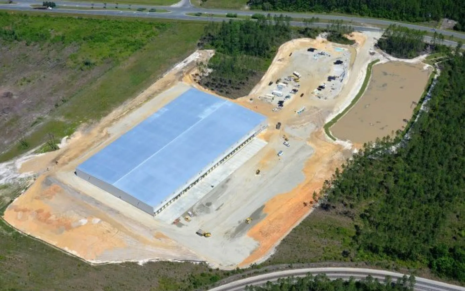 Aerial view of a large warehouse under construction surrounded by cleared land and forested areas.