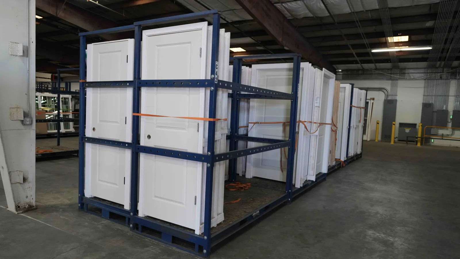 White interior doors stored upright on metal racks inside a warehouse with concrete floor and industrial lighting.