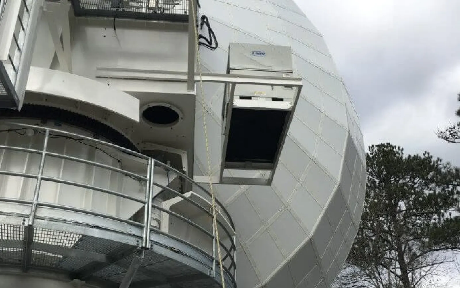 Large white radar dome structure with metal platform railing and trees in the background under cloudy sky