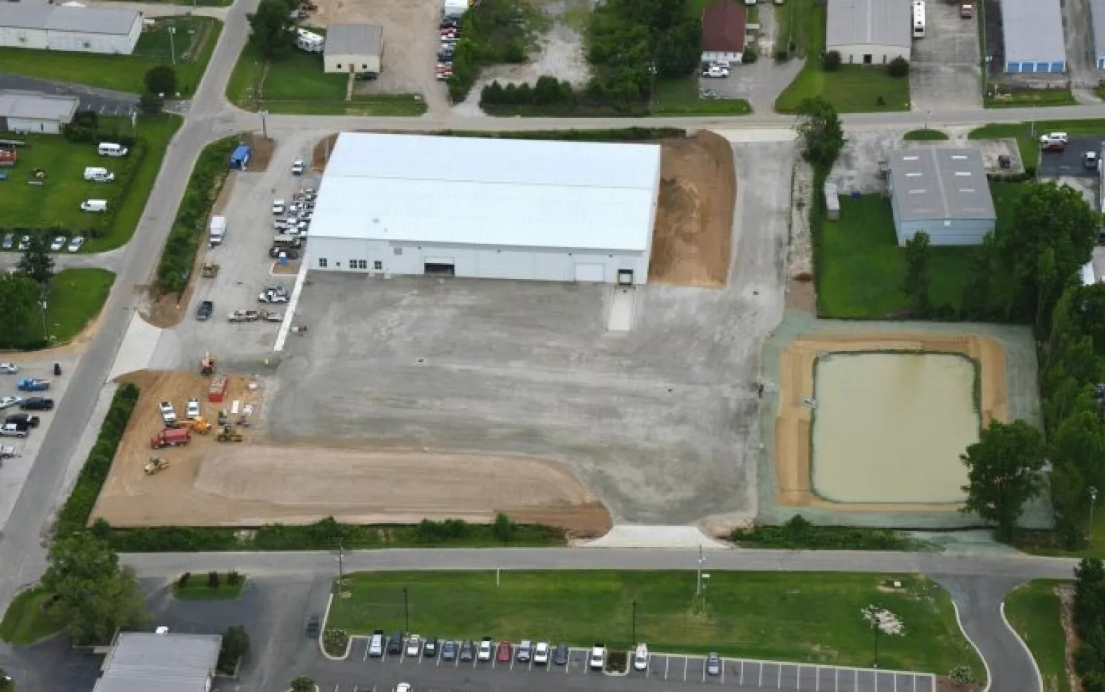 Aerial view of industrial warehouse with adjacent construction site and retention pond in a commercial area.