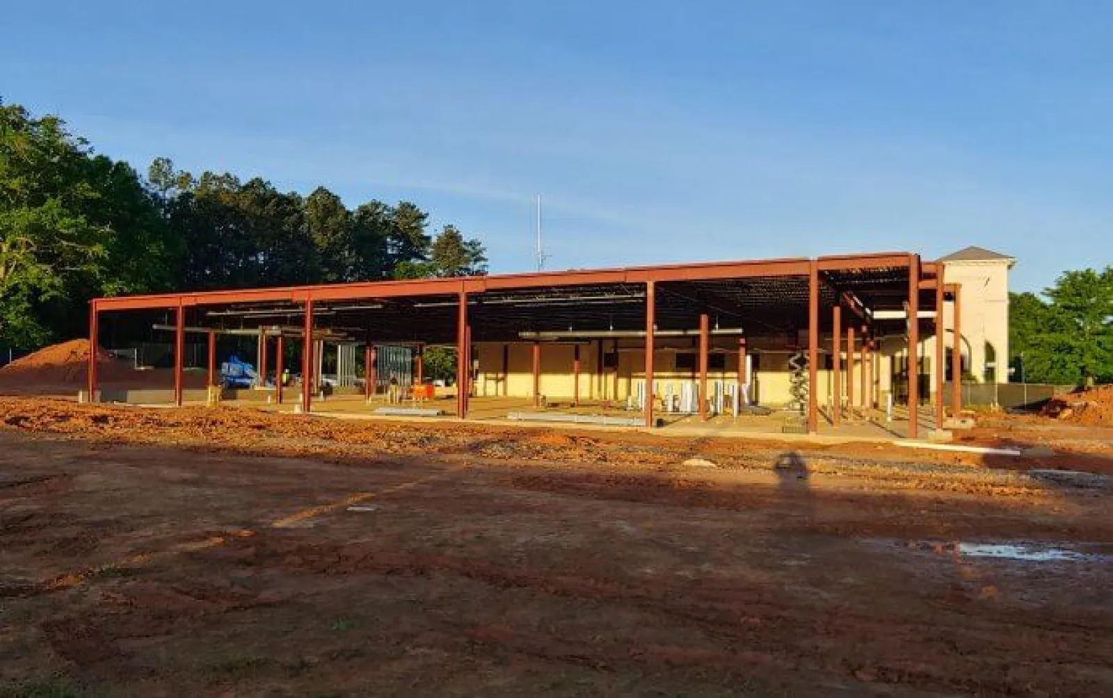 Steel framework of a building under construction on a cleared dirt site with trees and blue sky in the background