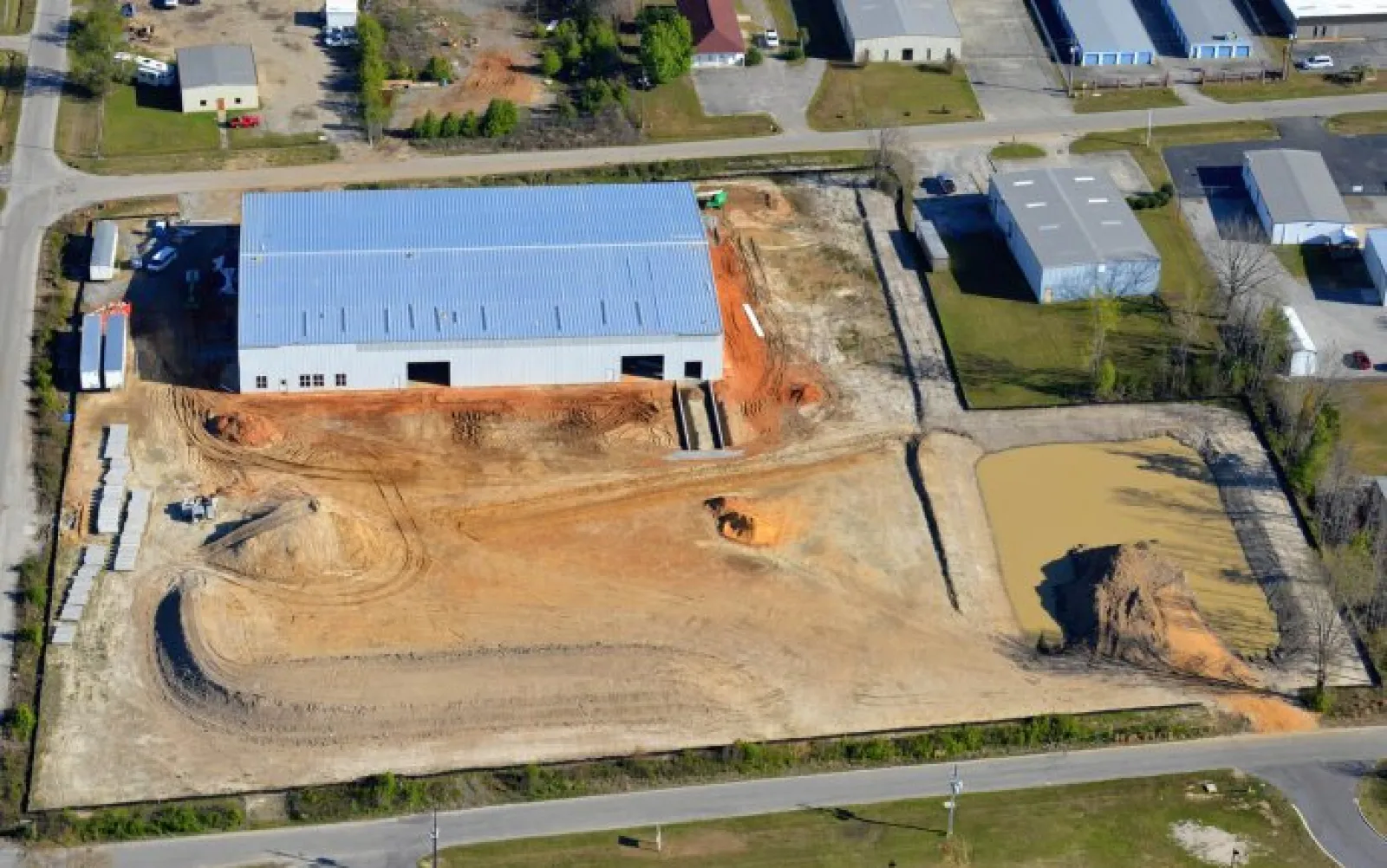 Aerial view of a construction site with a large warehouse, dirt roads, and a murky pond in an industrial area.