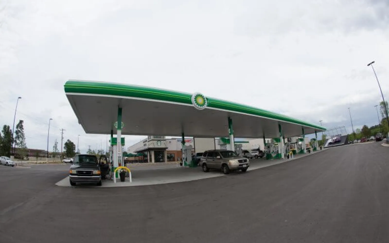 Wide view of a BP gas station with cars refueling under a large green and white canopy on a cloudy day.