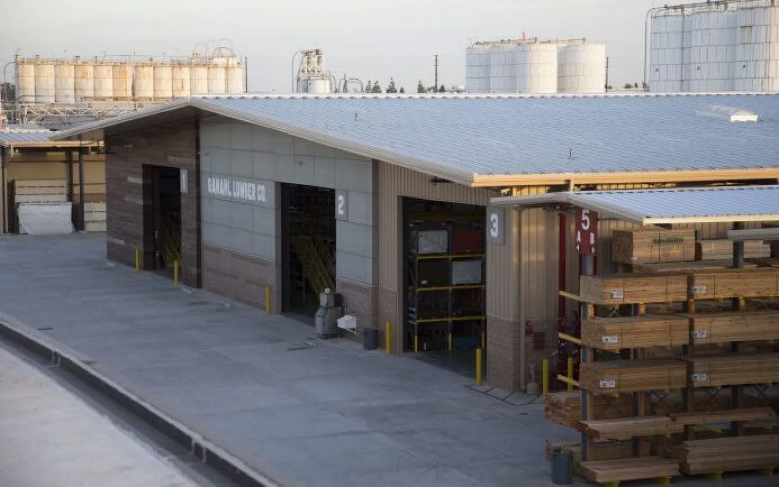 Outdoor lumber storage racks beside a warehouse building with industrial tanks in the background under a clear sky.