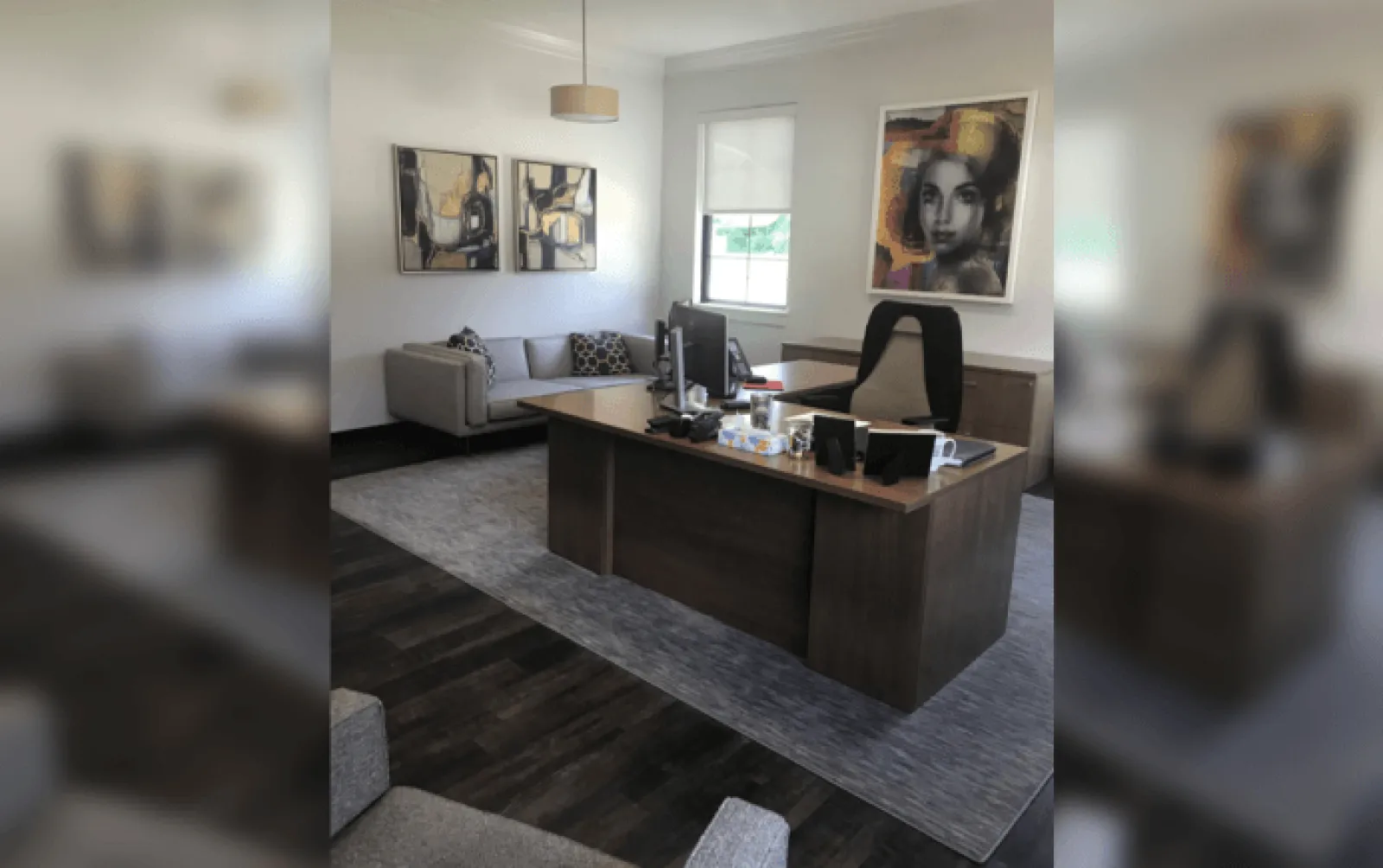 Modern office with wooden desk, black chair, couch, abstract art, and a large window with white walls.
