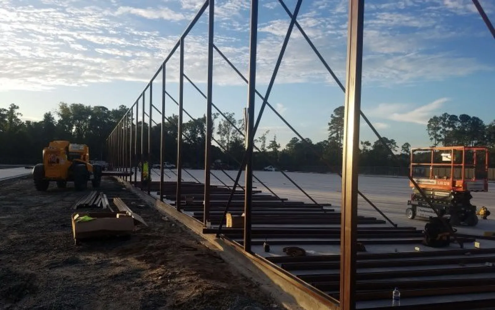 Steel framework under construction at a large outdoor site during early morning with construction equipment present