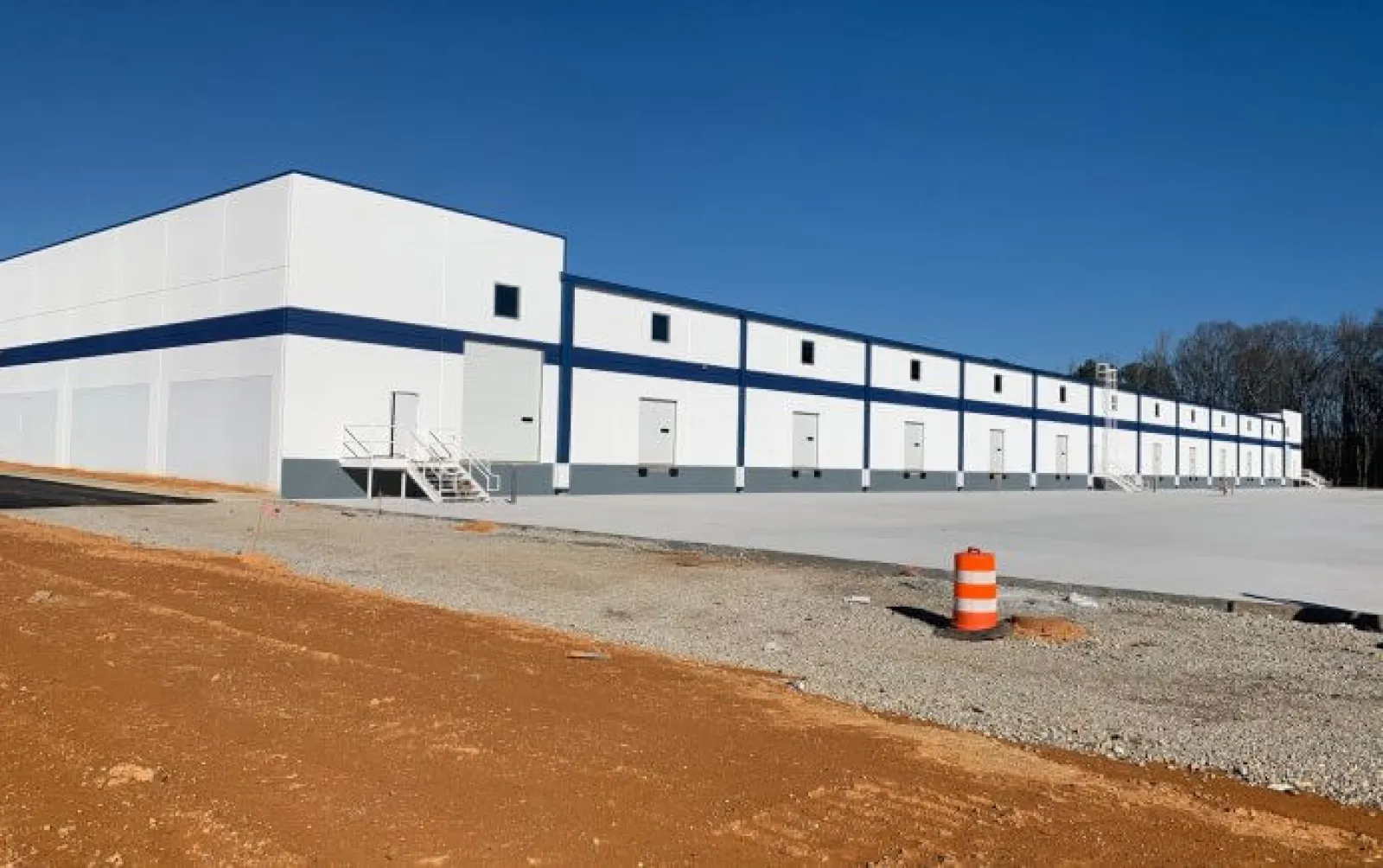 Large white industrial warehouse with a blue stripe under clear sunny sky, surrounded by dirt and gravel.
