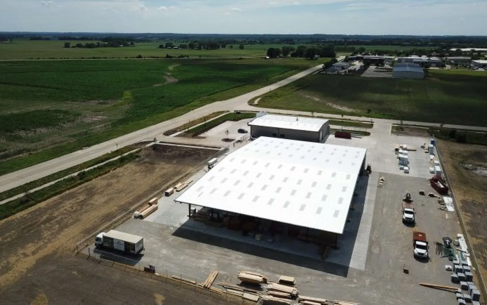 Aerial view of a large warehouse with white roof surrounded by farmland and vehicles on a sunny day