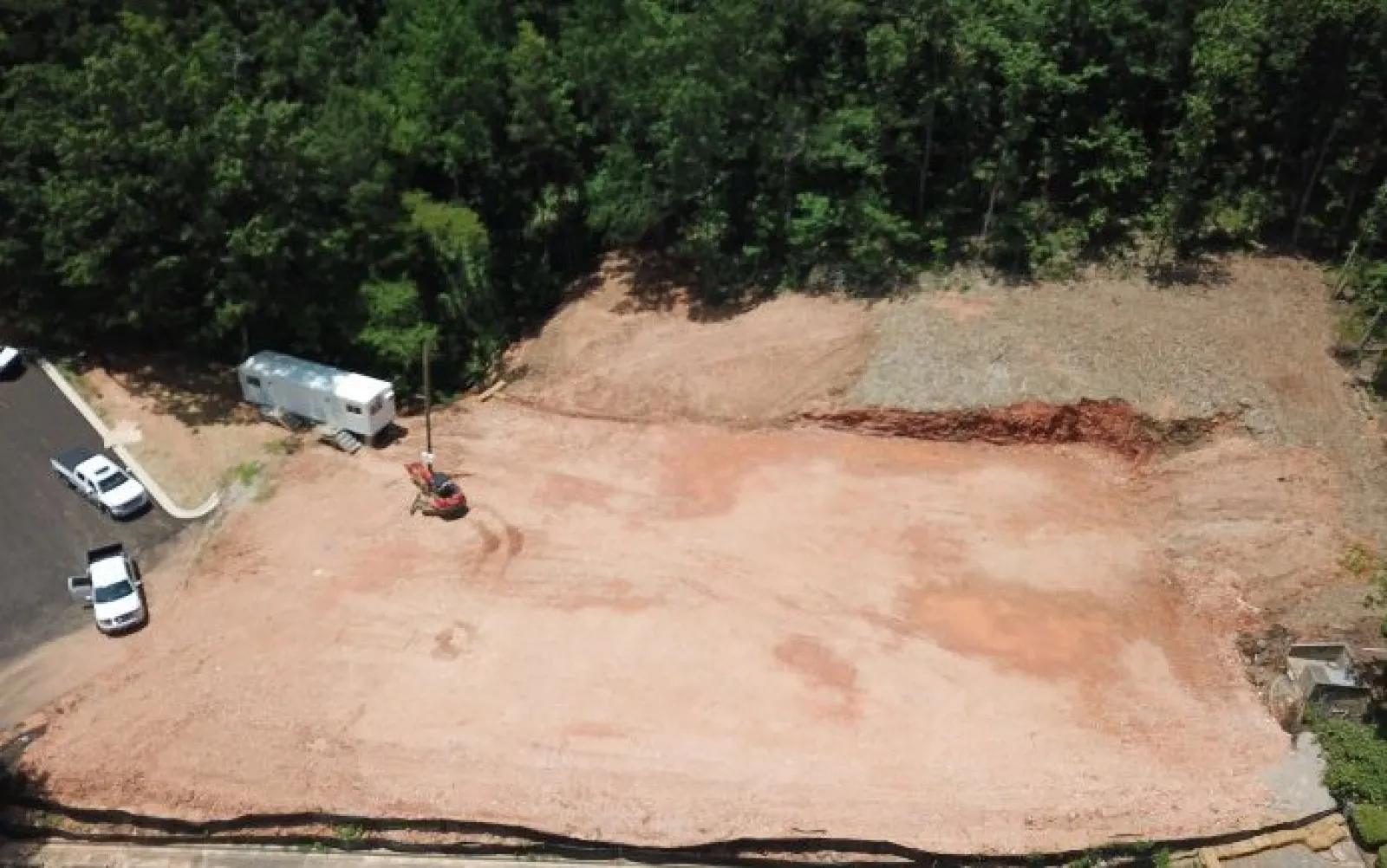 Aerial view of cleared construction site with dirt, parked vehicles, and surrounding trees in daylight.