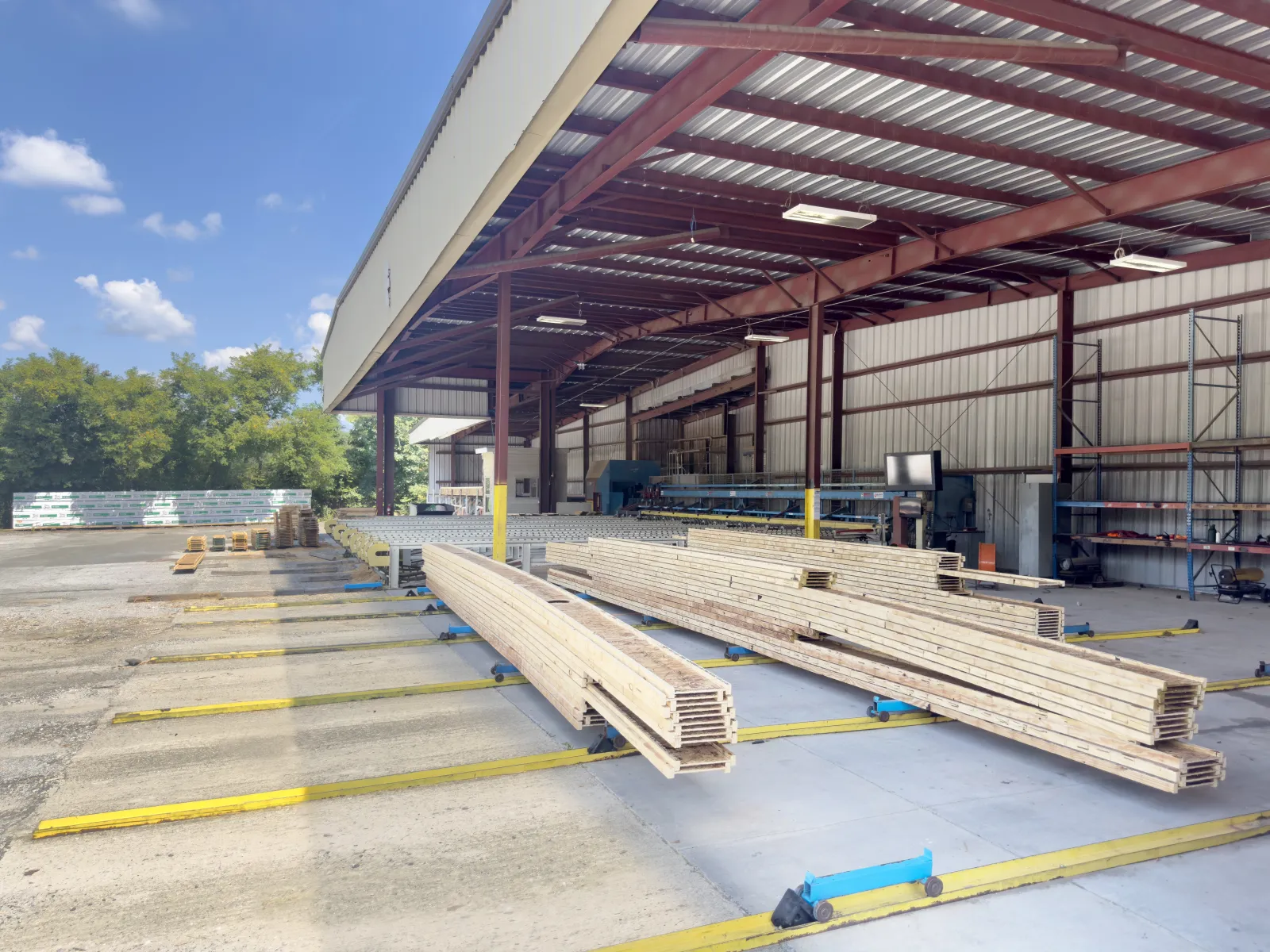 Stacked lumber under a metal roofed warehouse with clear sky and trees in the background