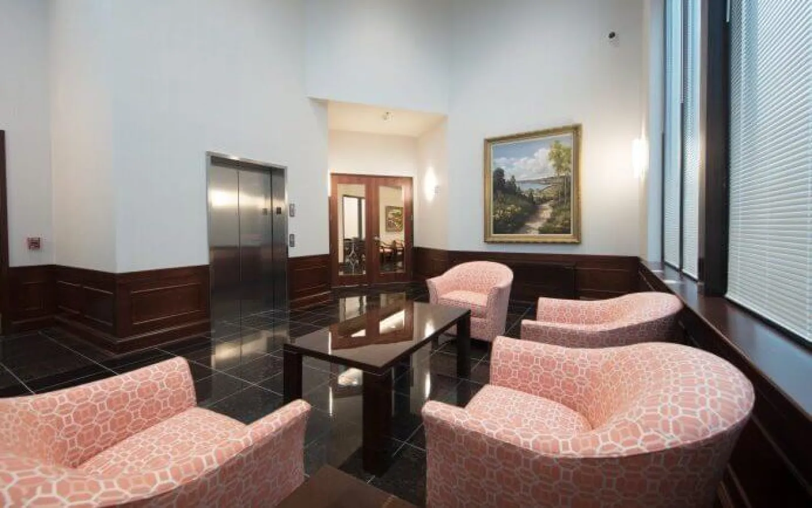 Lobby seating area with pink patterned chairs, glass coffee table, elevator, and framed landscape artwork.