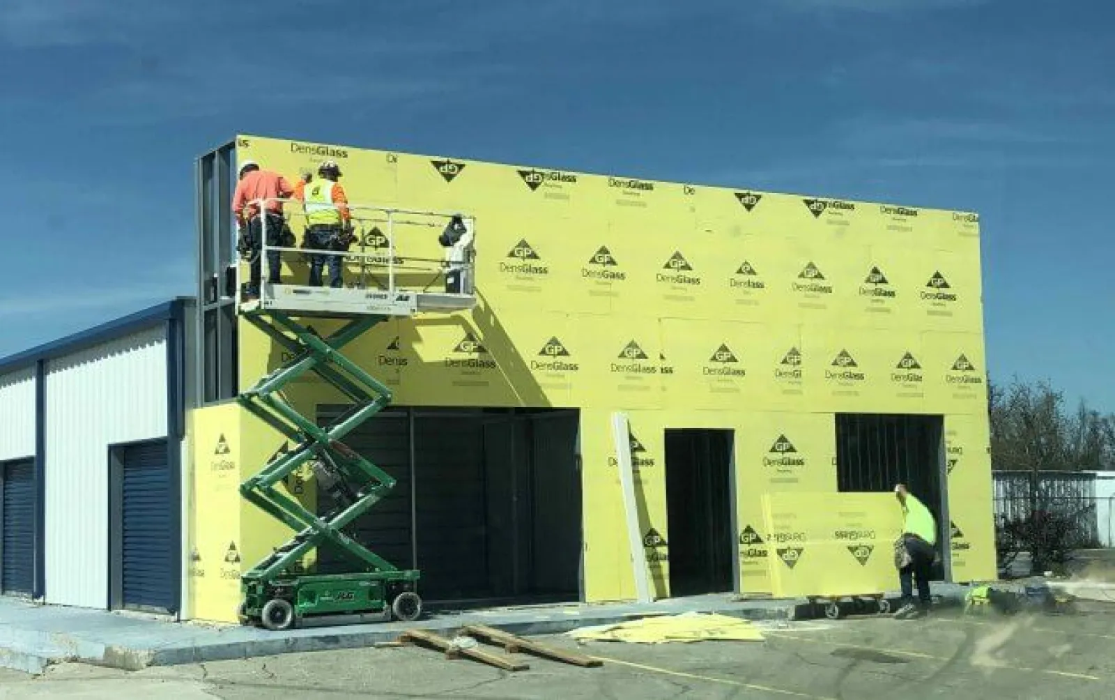 Construction workers install yellow DensGlass sheathing on a commercial building under clear blue sky.