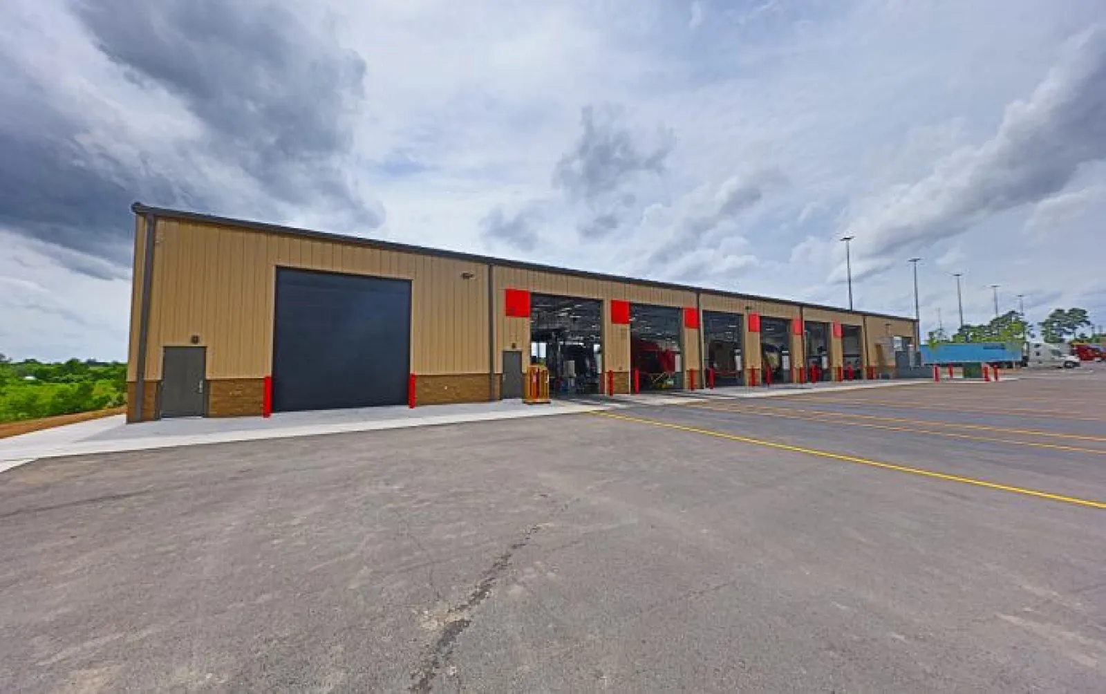 Wide view of a large industrial building with multiple open garage bays and a paved parking area under cloudy sky