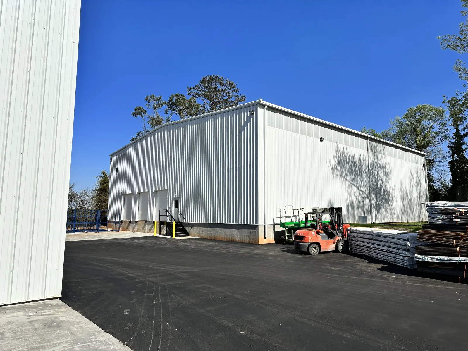 White industrial warehouse with forklifts and stacked pallets on asphalt under clear blue sky.