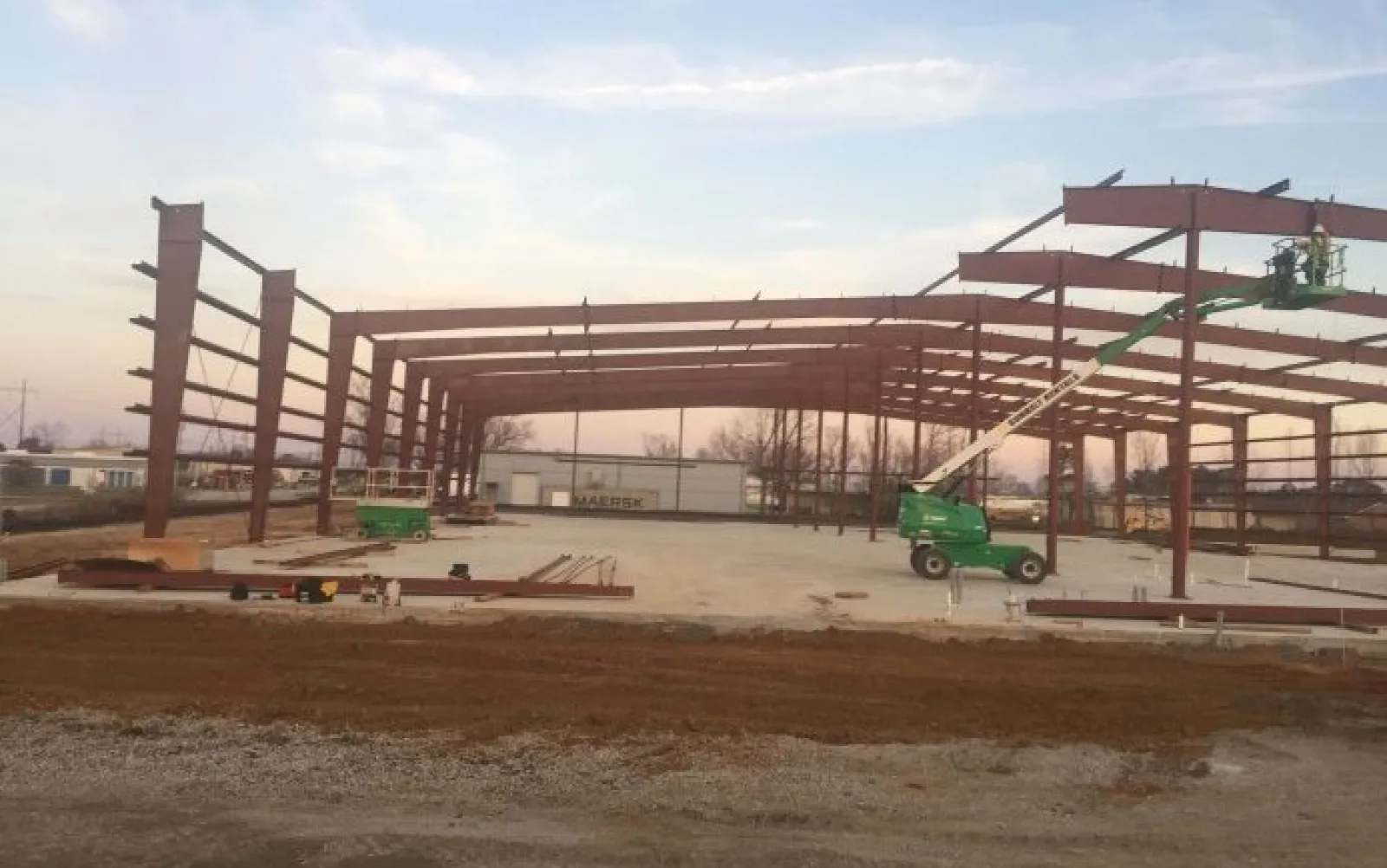 Steel frame structure under construction with a green boom lift and concrete foundation at dusk.