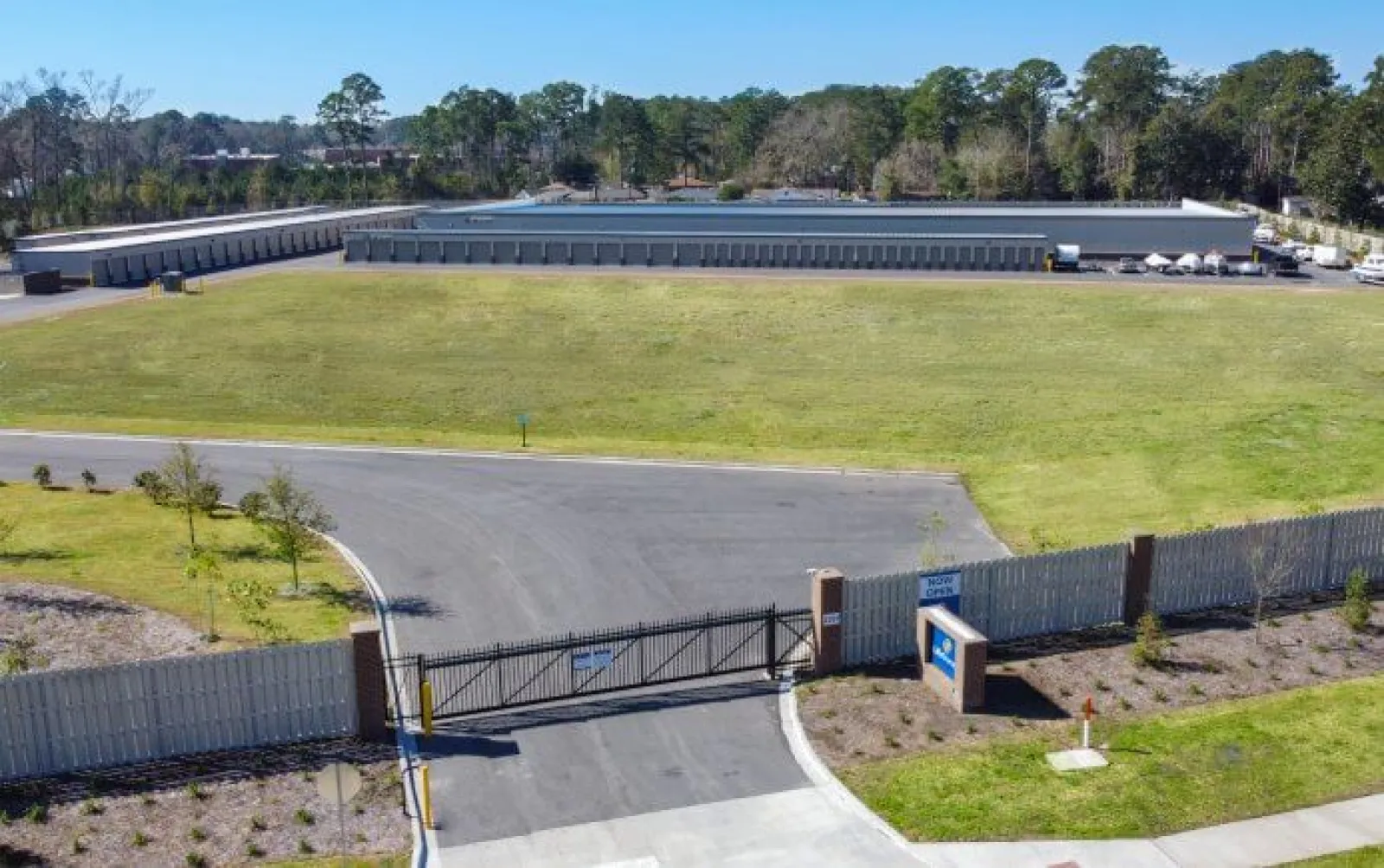 Aerial view of a gated self-storage facility with multiple storage units and large green lawn under clear sky.