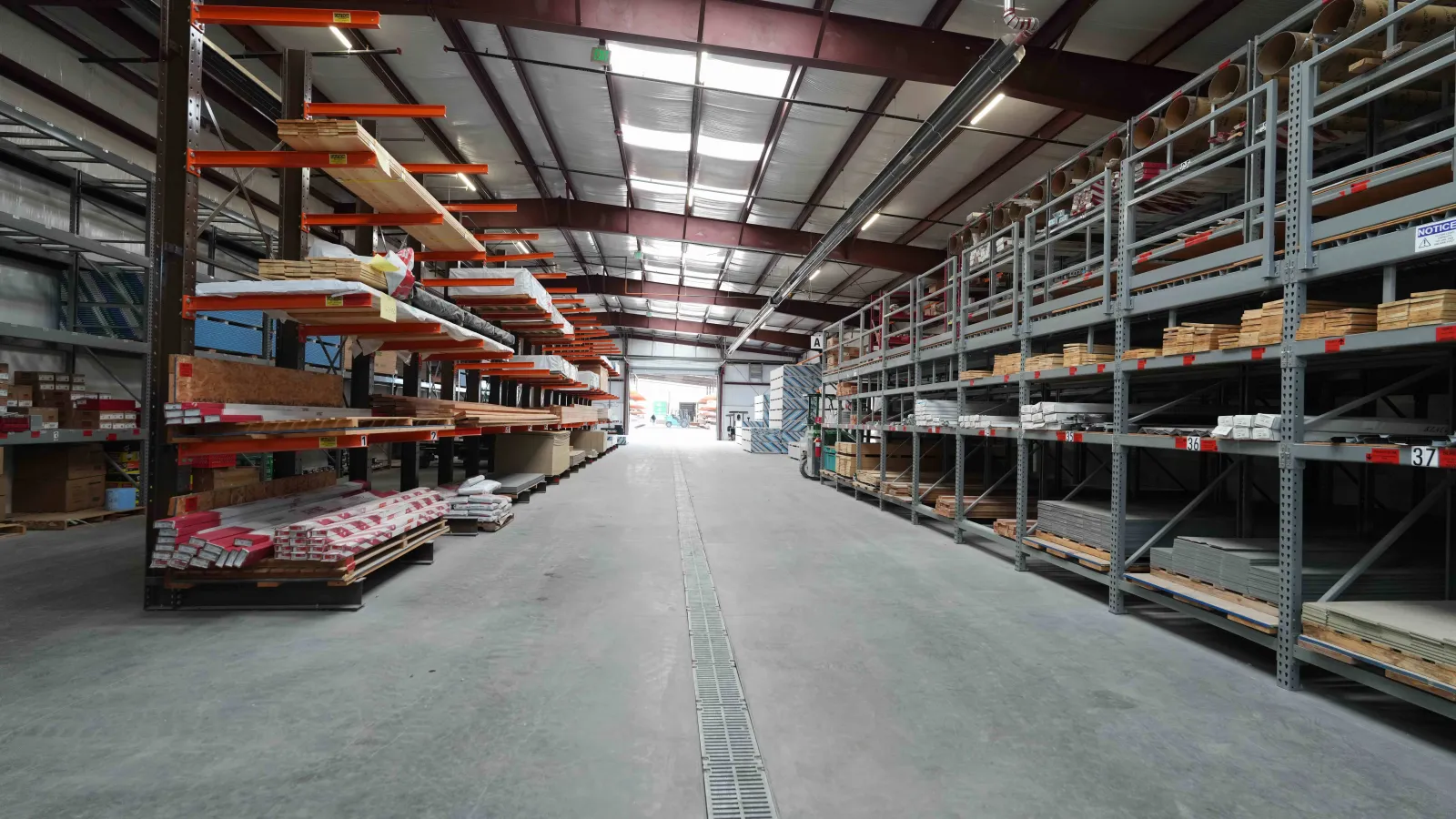 Interior view of a large warehouse with metal racks holding wood and building materials under a steel roof.