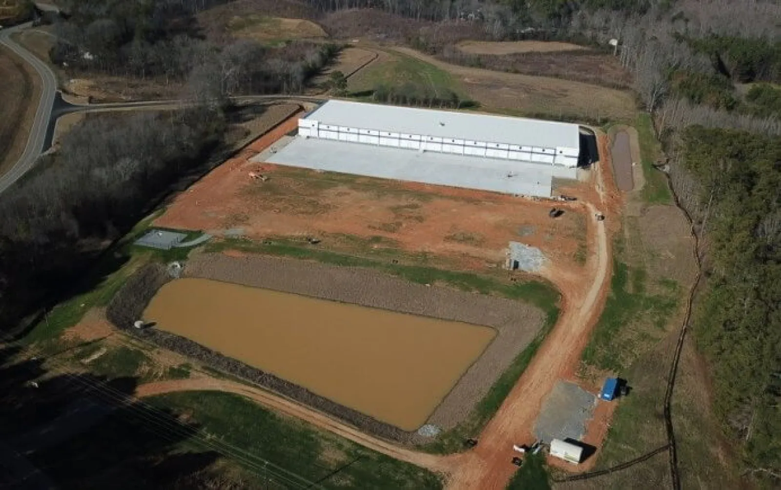 Aerial view of a large construction site with a white warehouse building near a muddy water retention pond.