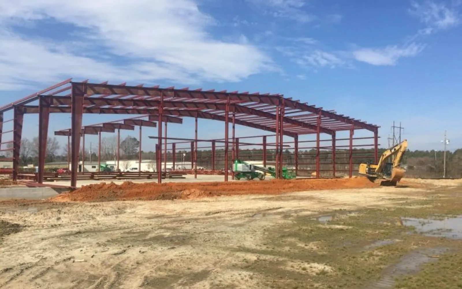 Steel frame structure under construction on a clear day with construction equipment and muddy ground in the foreground