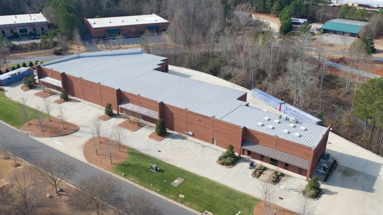 Aerial view of a large brick industrial building with a metal roof surrounded by parking areas and trees.