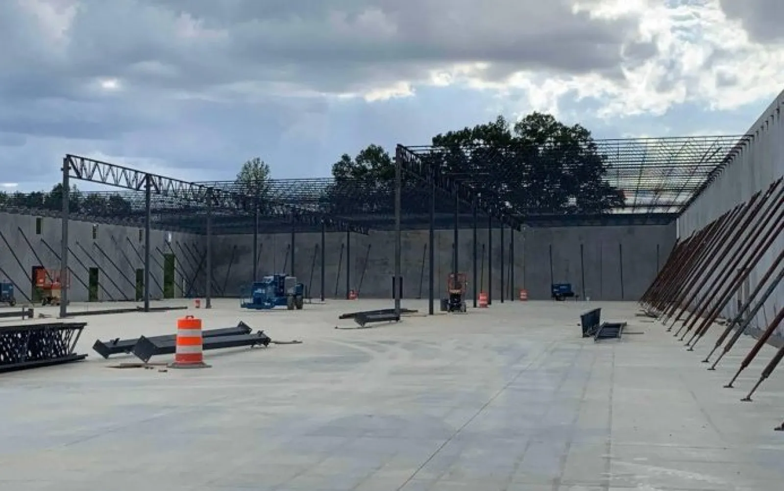 Construction site with concrete floor, steel framework, and orange traffic barrels under a cloudy sky.