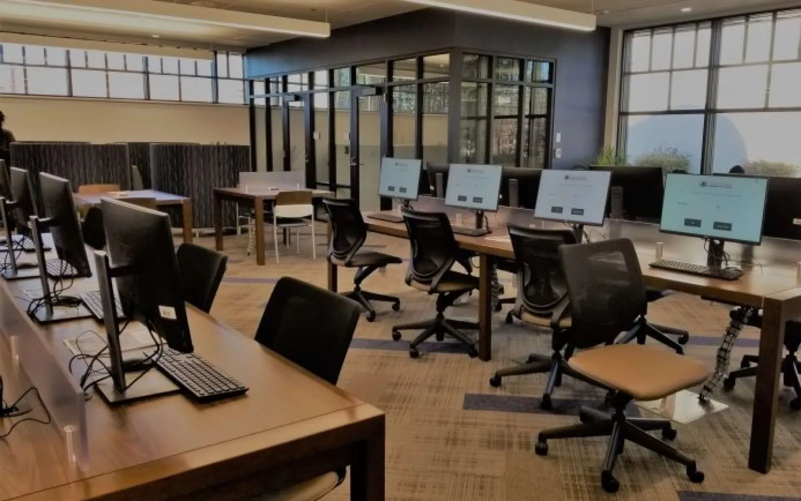 Modern office workspace with multiple computer stations, ergonomic chairs, and large windows providing natural light.