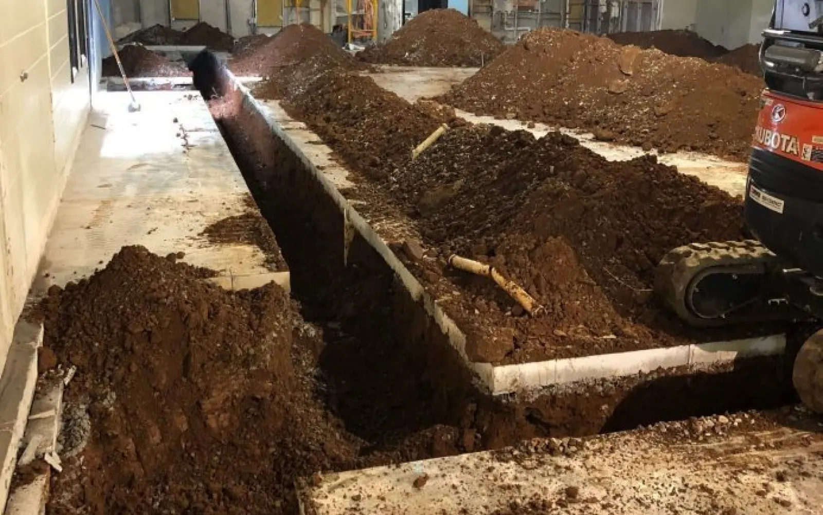 Indoor construction site with deep trenches dug in concrete floor and piles of soil around machinery.