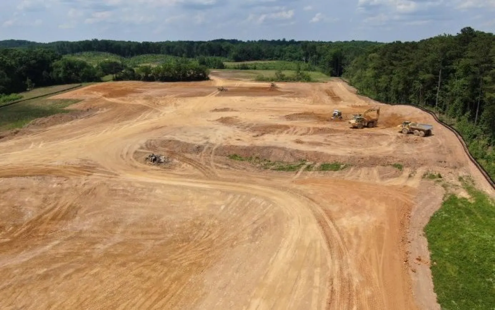Large construction site with dirt roads, heavy machinery, and forested background under partly cloudy sky