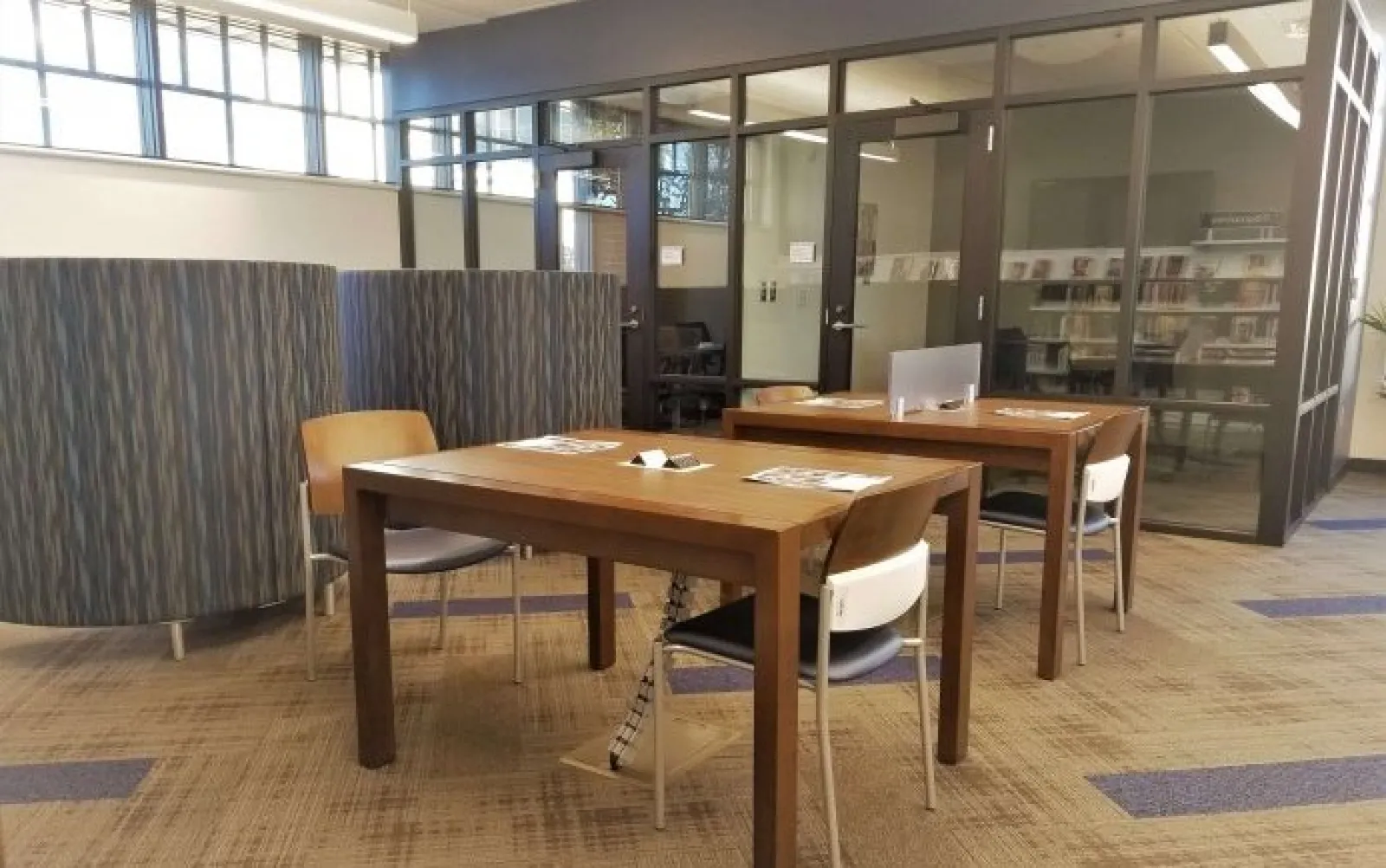 Modern study area with wooden tables, chairs, newspapers, and glass-walled office in a well-lit room.
