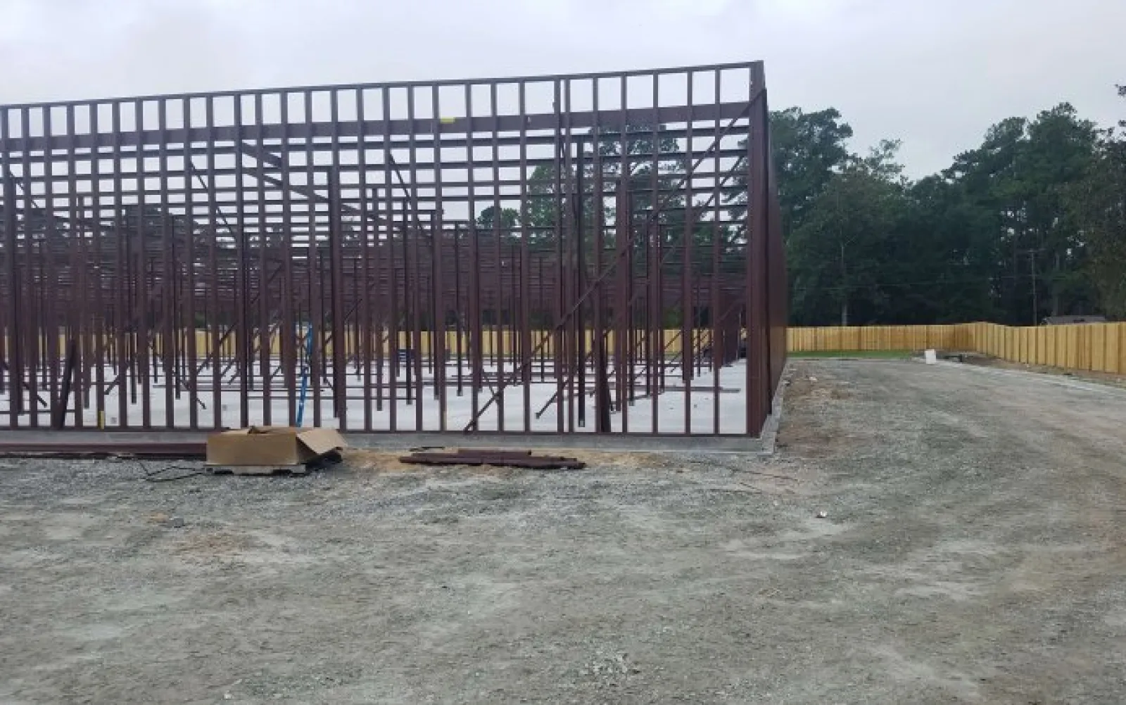 Steel framework of a building under construction on a cleared dirt site with a wooden fence and trees in the background.