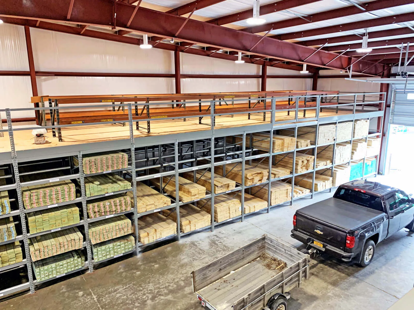 Warehouse interior with stacked lumber on metal racks and a black pickup truck beside a wooden trailer under a metal roof