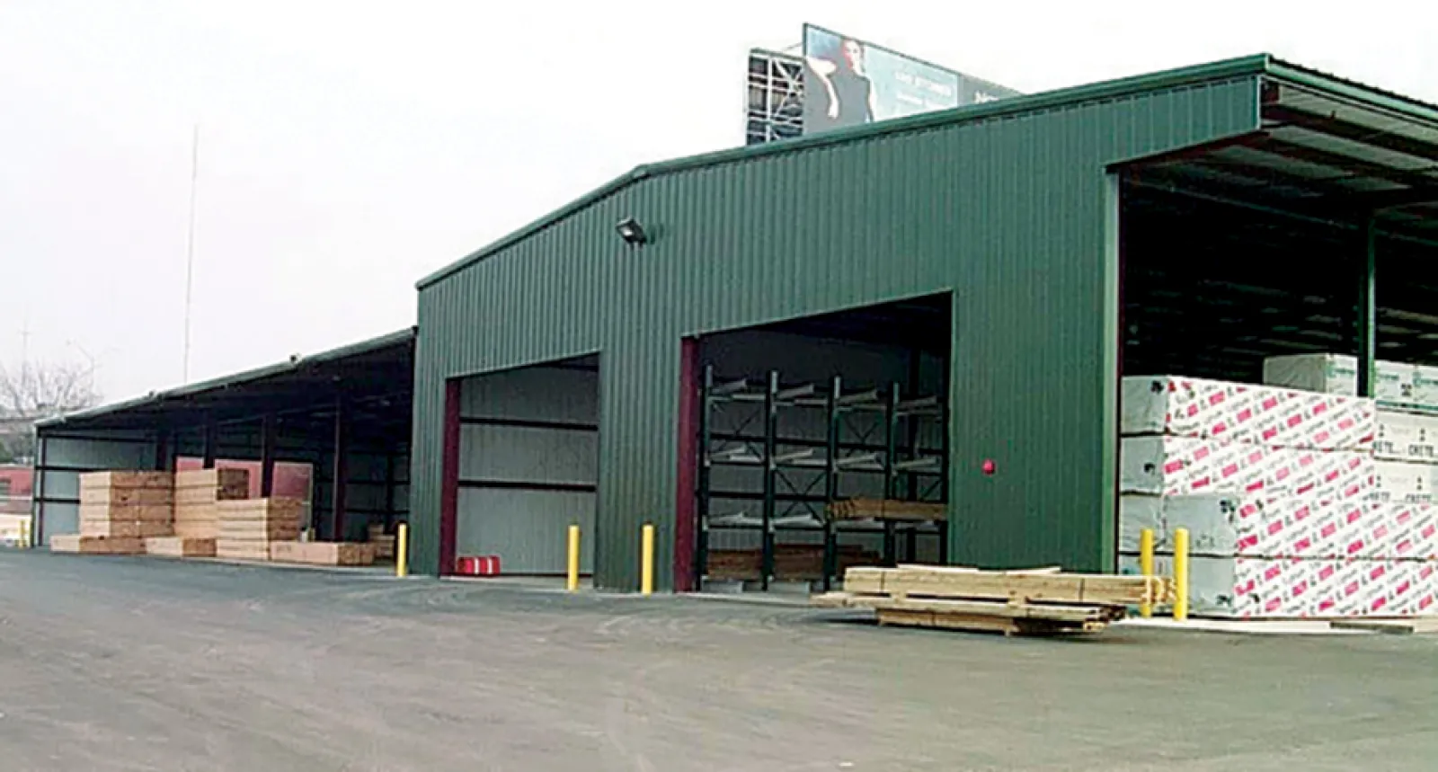 Large green warehouse storing various wood planks and construction materials with open bays.