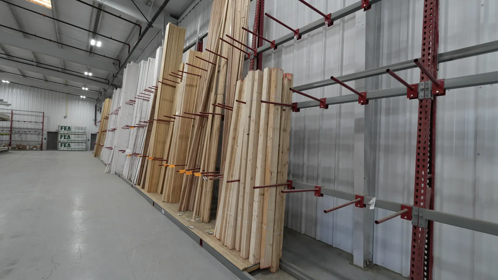 Stacks of wooden planks stored vertically on red metal racks inside a warehouse with high ceilings.