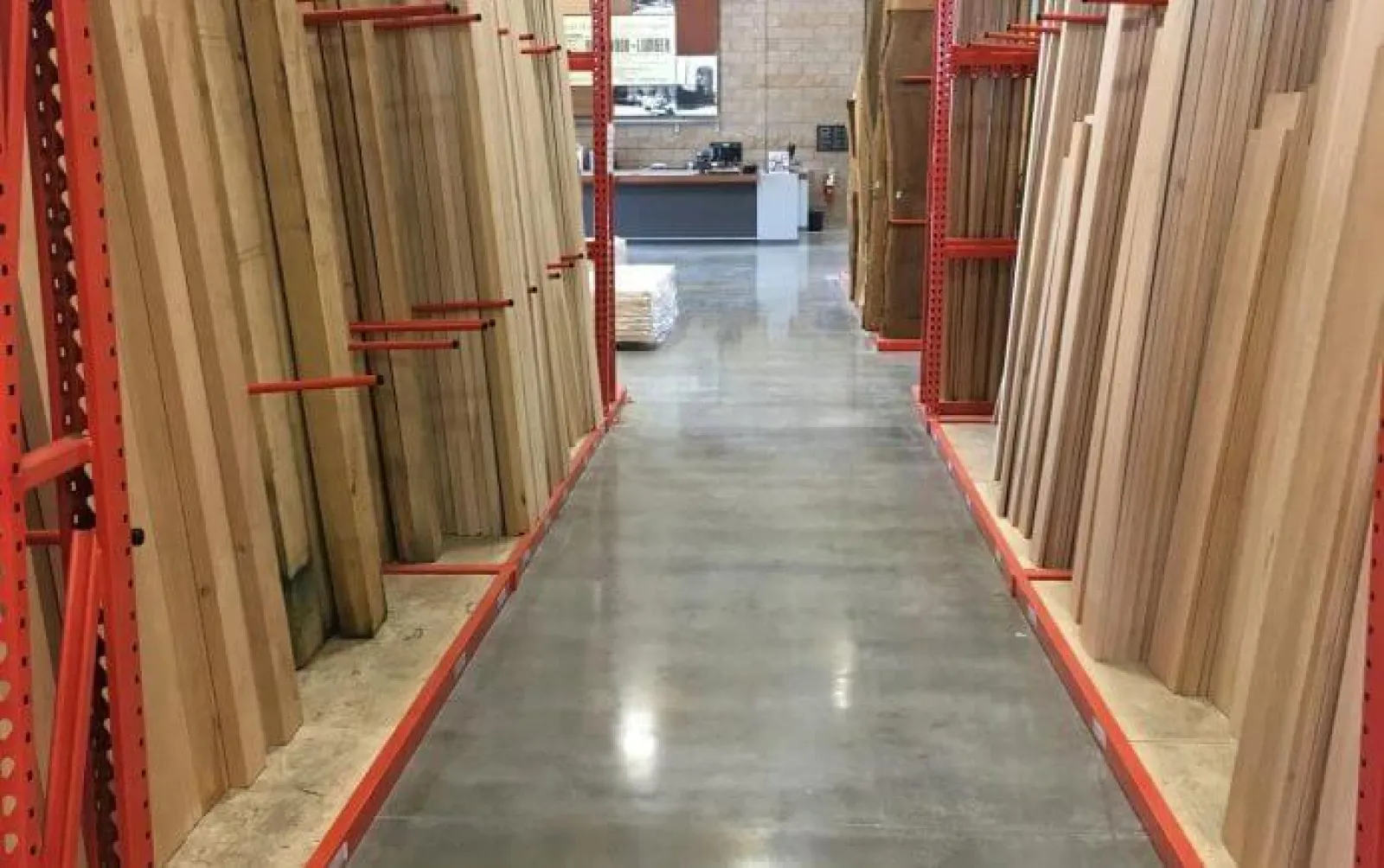 Aisle in a hardware store with stacks of wooden boards organized on red metal racks on both sides.